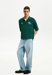 Grön polo skjorta med krage, med vita logodetaljer. Löst sittande ljusblåjeans och svarta skor fulländar outfiten.