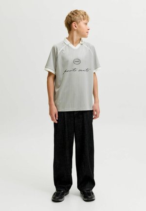 Blond chłopiec stoi zwrócony w prawo, ubrany w szarą, prążkowaną koszulkę „porto santo”, czarne szerokie spodnie i czarne trampki na białym tle.