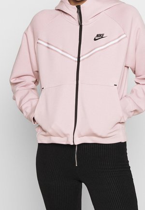 Veste à capuche zippée rose avec logo Nike noir, rayures blanches formant un design en chevron sur la poitrine, et poches avec une texture lisse.