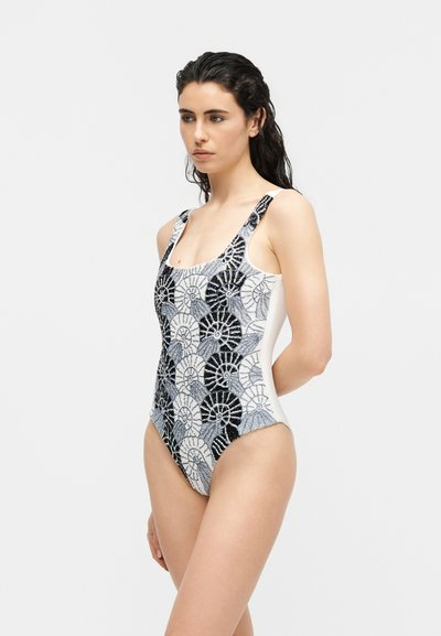 Femme aux cheveux foncés et mouillés, portant un maillot de bain une pièce à motifs coquillages noir, blanc et gris, se tenant les bras derrière le dos.