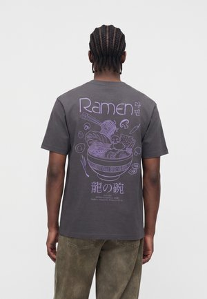 Uomo con capelli intrecciati che indossa una maglietta grigio scuro con un'illustrazione di una ciotola di ramen viola e testo in inglese, coreano e giapponese sulla schiena.