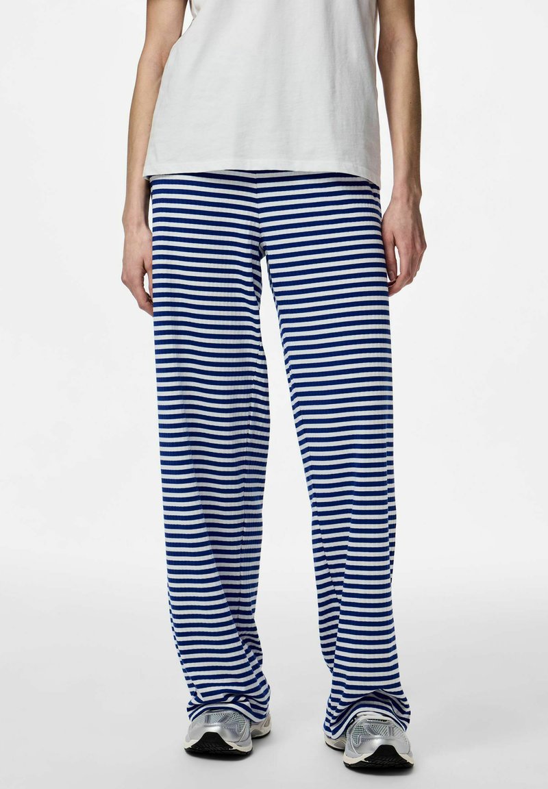 Inimene kannab lahtisi sini-valgeid horisontaalse triibuga pükse, valget tuba ja hõbedasi tossusid, seisab valge tausta ees.