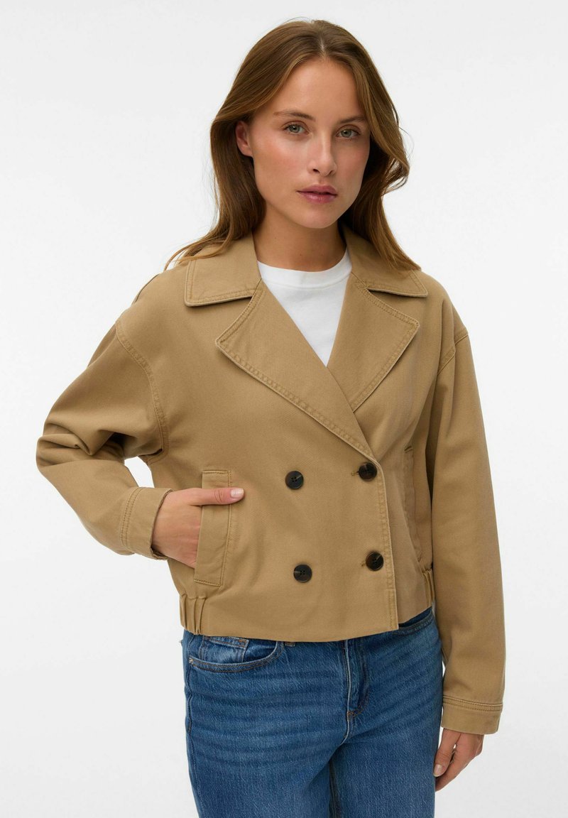 Femme portant une veste beige à double boutonnage avec des boutons noirs et un jean bleu, main dans la poche, sur un fond blanc uni.