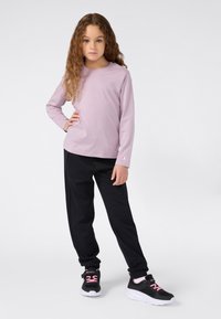 Ein junges Mädchen mit langen, lockigen Haaren, das ein lila Langarmshirt, schwarze Hosen und schwarze Turnschuhe mit pinken Schnürsenkeln trägt, steht mit einer Hand auf der Hüfte.