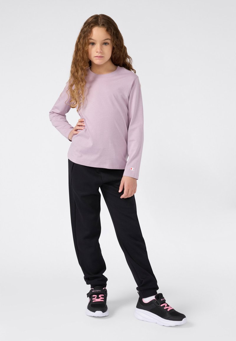 Ein junges Mädchen mit langen, lockigen Haaren, das ein lila Langarmshirt, schwarze Hosen und schwarze Turnschuhe mit pinken Schnürsenkeln trägt, steht mit einer Hand auf der Hüfte.
