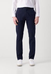 Person som bär marinblå rak passform byxor, vit skjorta med knappar och vita sneakers står mot en neutral bakgrund.