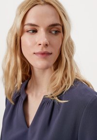 Navyblauwe blouse met een V-hals, zachte textuur en subtiele plooien bij de schouders, gedragen met casual, losse golven in het haar.