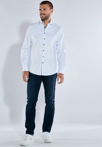 Mann in einem weißen Hemd mit Knöpfen, dunklen blauen Jeans und weißen Sneakers, der vor einem schlichten hellgrauen Hintergrund steht.