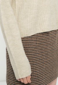 Beige neulepaita, jossa on ribbieritys, yhdistetty ruskeaan houndstooth-hameeseen. Neulepaidassa on hieman löysä istuvuus ja risaiset hihansuut.