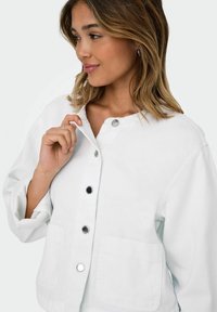 Veste en denim blanc avec un col rond, des fermetures à pression à l'avant et deux grandes poches. Dotée de manches longues et d'une coupe décontractée.