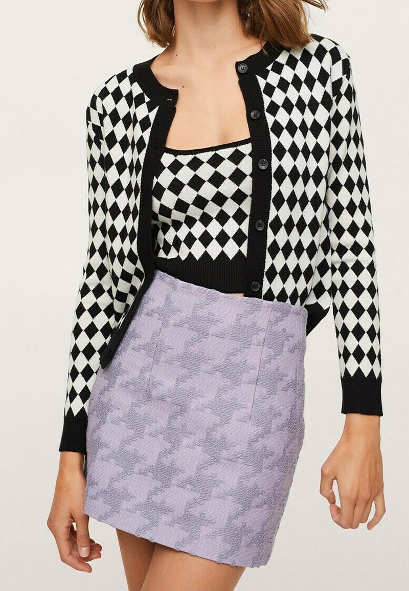 Femme portant un cardigan et un haut à motif losange noir et blanc avec une mini-jupe lavande texturée.