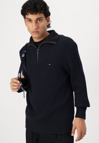 Marineblauer, gerippter Pullover mit halbem Reißverschlusskragen, mit strukturierten Details, einem kleinen Logo und enganliegenden Bündchen. Das Modell hält eine Sonnenbrille.