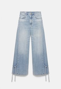 Jean bleu clair à jambes larges avec poches avant et liens réglables près de l'ourlet sur les deux jambes, posé à plat sur un fond blanc.