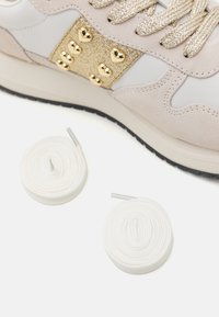 Beige és fehér felsőrészű sneakers, arany csillám díszítéssel, szív alakú szegecsekkel, mellette két fehér cipőfűző.