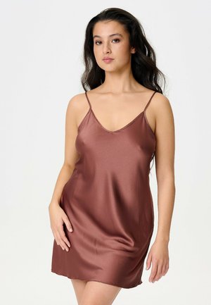 Femme aux longs cheveux foncés portant une robe nuisette en satin marron à fines bretelles, debout devant un fond clair uni.