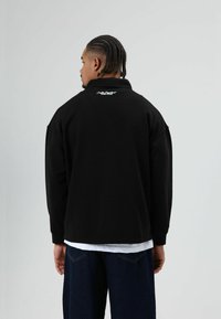 Zwarte pullover jas met een hoge kraag, losse pasvorm en geborduurd logo op de achterkant. Draagt een wit overhemd eronder en donkere spijkerbroek.