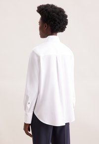 White button-up shirt with long sleeves, a back pleat, and rounded hem. Features buttoned cuffs with a black embroidered emblem.