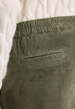 Pantalon en velours côtelé olive avec une surface texturée, présentant un design en côtes et une petite poche arrière. Ceinture élastique en haut.