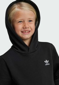 Čierna mikina s kapucňou s mäkkou textúrou a bielym logom Adidas na ľavej strane hrudníka. Kapucňa je plne integrovaná a zaoblená.
