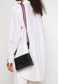 Donna che indossa una camicia bianca larga e pantaloni, con una borsa a tracolla nera dotata di un logo circolare e una tracolla a strisce rossa, bianca e nera.