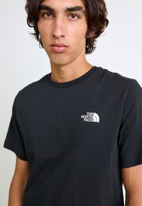 Sort bomulds t-shirt med rund halsudskæring og korte ærmer, som tydeligt viser et hvidt logo på venstre brystområde.