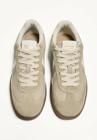 Beige ruskinds sneakers med en glat finish, prydet med hvide detaljer, flade snørebånd og en rund tå, sat på en struktureret brun gummisål.