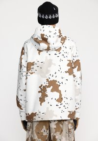 Witte camouflagejas met bruine en zwarte patronen, met een capuchon en elastische manchetten, gecombineerd met beige camouflagebroeken.