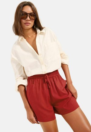 Kobieta nosząca za dużą kremową koszulę zapinaną na guziki, włożoną w czerwone szorty z wysokim stanem i sznurkiem, z brązowymi prostokątnymi okularami przeciwsłonecznymi i kolczykami.