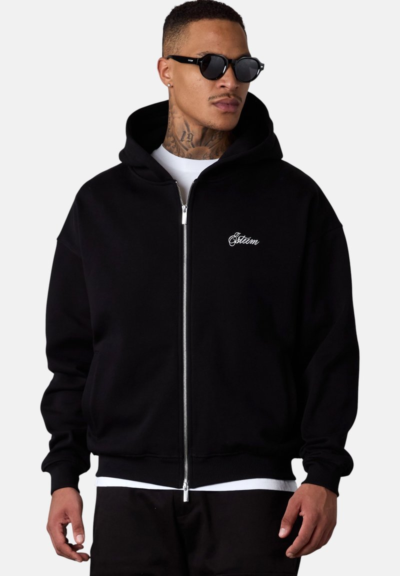 Haut de hoodie noir zippé avec une capuche structurée, des poignets à côtes et un ourlet. Présente un logo brodé sur la poitrine, fabriqué en tissu doux.
