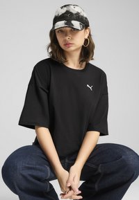 Čierne voľné tričko s bielym logom Puma; doplnené šedou šnúrovanou čiapkou. Model sedí so zloženými rukami, na ušiach má veľké kruhové náušnice.