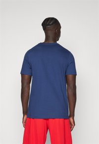 T-shirt in cotone blu navy con maniche corte e collo girocollo, vista da dietro, abbinata a pantaloni corti rossi. Texture liscia e design semplice.
