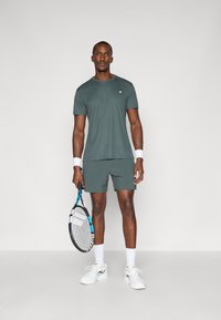 Ανδρική αθλητική ενδυμασία με σκούρο πράσινο μπλουζάκι και αντίστοιχα σορτς. Κρατάει ένα ρακέτα τένις και φοράει άσπρες κάλτσες και παπούτσια. Απαλή ύφανση, ελάχιστες λεπτομέρειες.