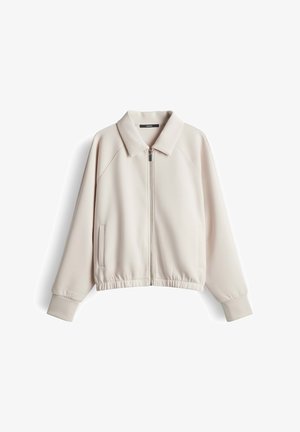 Beige lange mouw jas met rits en kraag, elastische zoom, zijzakken en geribde manchetten, plat liggend op een witte achtergrond.