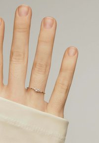 Anillo de oro rosa con tres pequeños acentos similares a diamantes, llevado en una mano con un acabado de uñas neutro, contra un fondo claro.