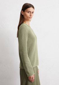 Maglione lungo a maniche lunghe di un verde chiaro texturizzato con scollatura rotonda, vestibilità rilassata e polsini a coste, abbinato a una gonna verde.