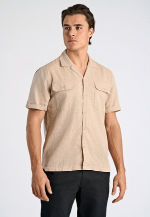 Chemise à manches courtes, boutonnée, de couleur beige clair, avec deux poches poitrine et une texture subtile. Manches retroussées et coupe décontractée.