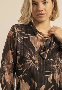 Blouse à motif floral en tissu léger et transparent ; présente un lien au niveau du col et des tons terreux de marron, beige et noir.