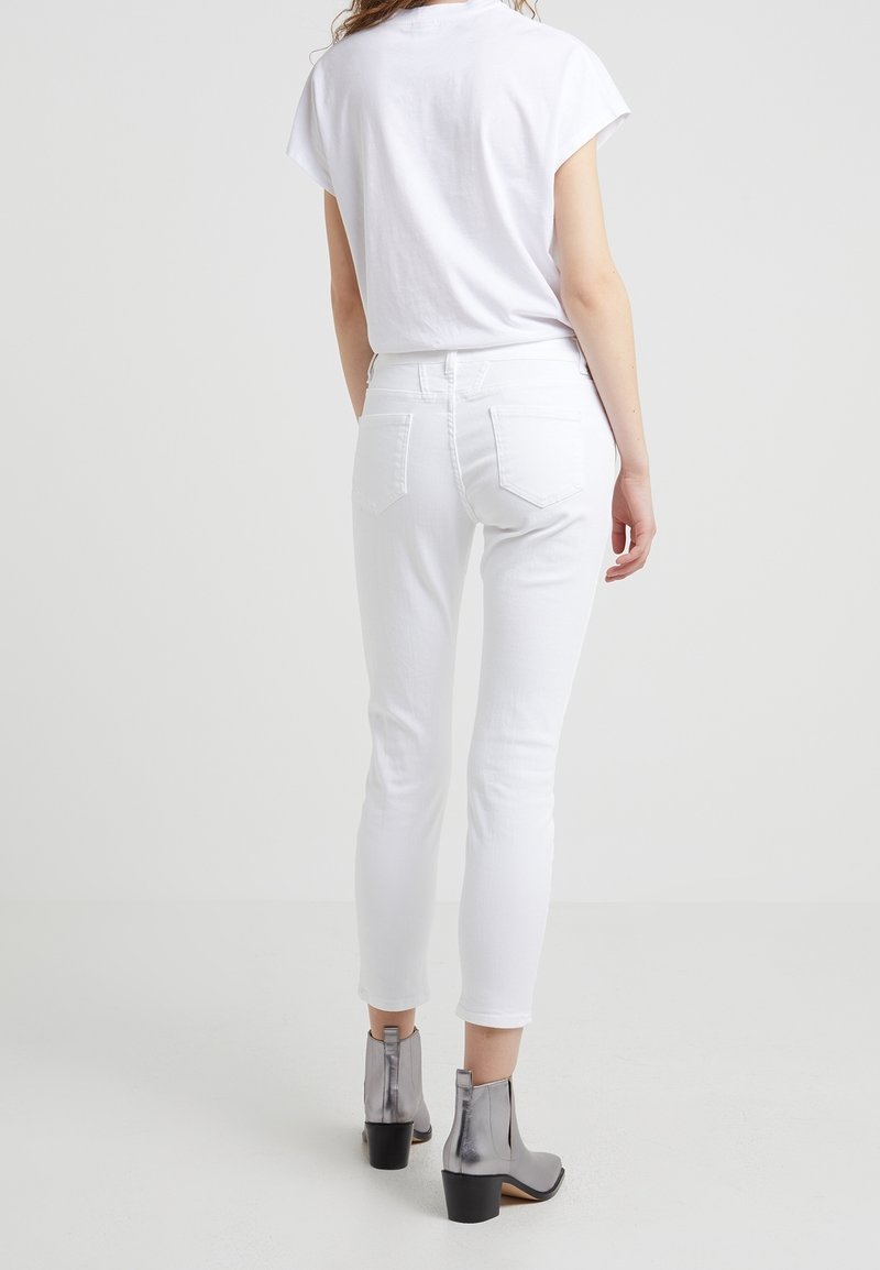 Personne portant un T-shirt blanc à manches courtes, un pantalon blanc ajusté jusqu'à la cheville et des bottines argentées, debout devant un fond clair uni.