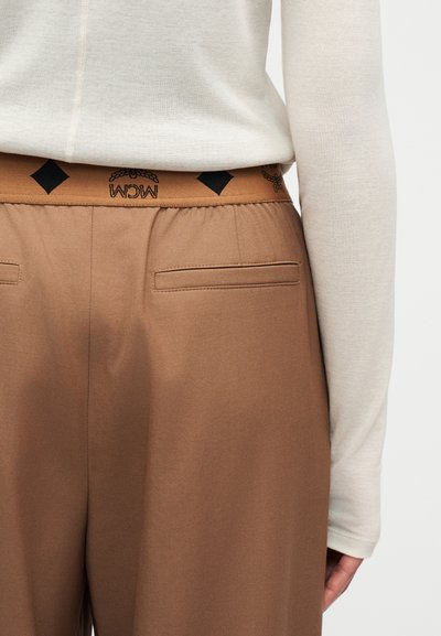 Gros plan sur une personne portant un haut beige à manches longues rentré dans un pantalon marron avec une taille élastique ornée de motifs de losanges noirs et de feuilles de laurier.