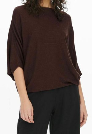 Femme portant un haut en maille côtelée marron foncé ample à manches trois-quarts et un pantalon noir, debout devant un fond blanc.