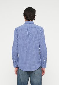 Homme vu de dos portant une chemise à manches longues à carreaux bleu et blanc et un jean en denim bleu sur un fond uni.