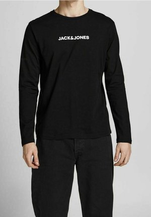 Personne portant un haut à manches longues noir avec le logo "JACK&JONES" en blanc centré sur la poitrine, associé à un pantalon noir.