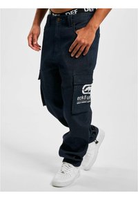 Dark blue cargo pants made of denim, featuring large side pockets, a white logo on the thigh, and a ribbed waistband with lettering.