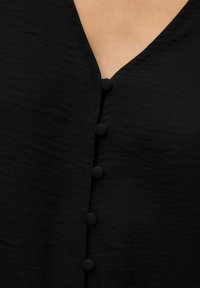 Blusa nera con scollatura a V e sei bottoni rivestiti in tessuto lungo la parte anteriore, con una finitura texturizzata.
