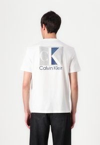 T-shirt en coton blanc avec une grande impression graphique bleue et grise portant le texte "CK" et "Calvin Klein" au dos. Manches courtes et col rond.
