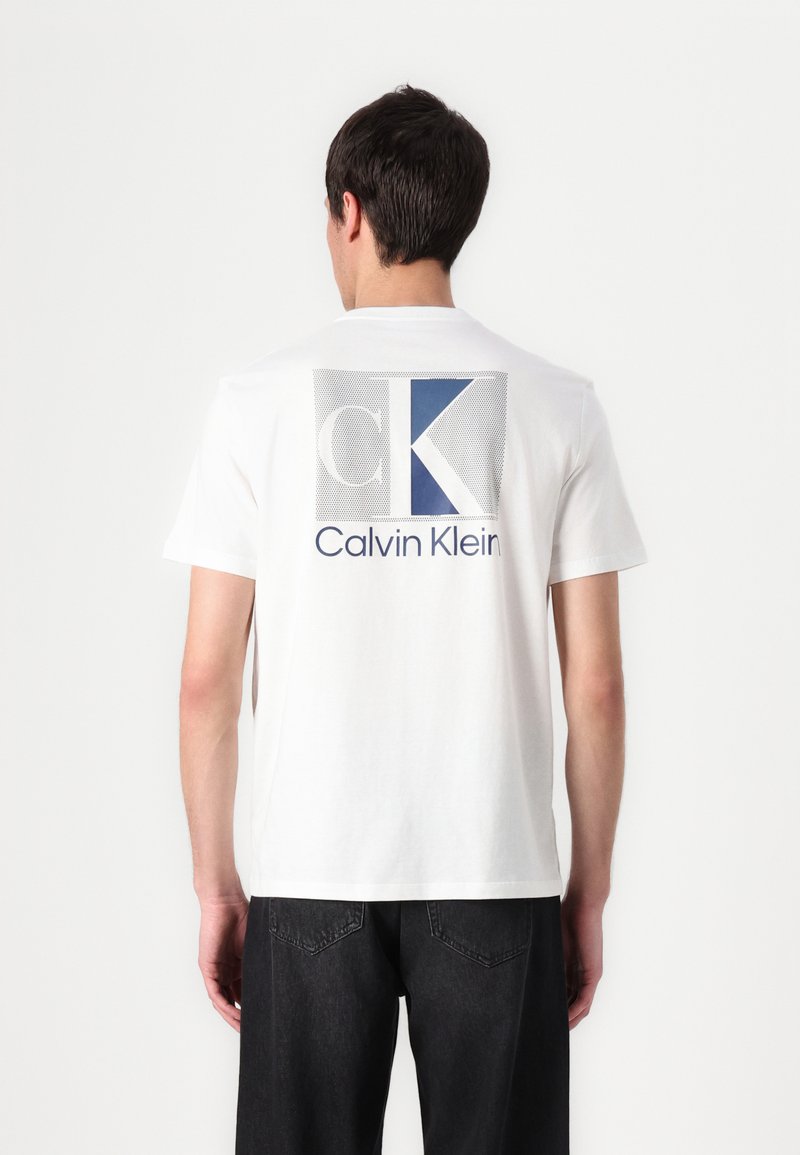 T-shirt en coton blanc avec une grande impression graphique bleue et grise portant le texte "CK" et "Calvin Klein" au dos. Manches courtes et col rond.