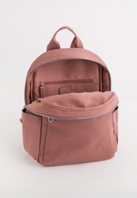 Mochila de tela rosa con textura suave, compartimento principal con cremallera, bolsillo frontal y asa de transporte. El interior incluye una pequeña área con cremallera.