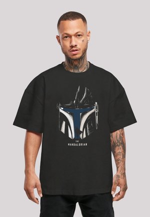 Sort t-shirt med et grafisk design af en Mandalorian-hjelm i hvid og blå. Designet inkluderer teksten "THE MANDALORIAN" nedenfor.