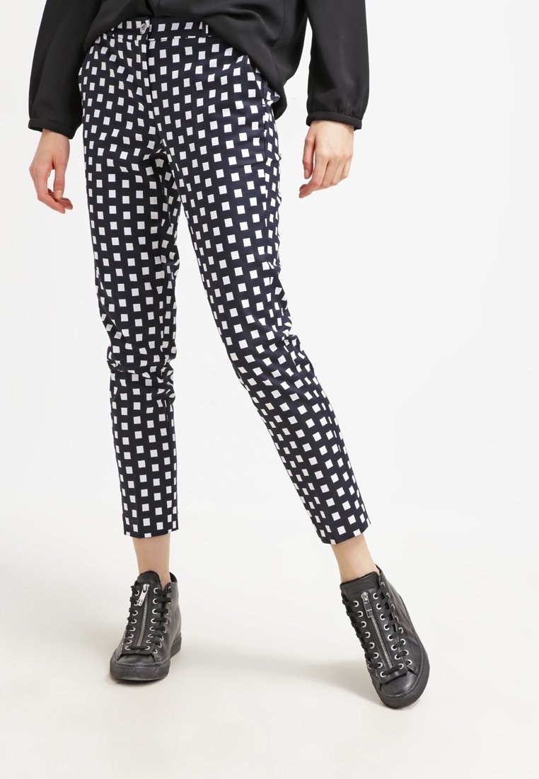 Personne portant un pantalon à carreaux noir et blanc, une veste noire et des baskets montantes noires, debout les jambes écartées.