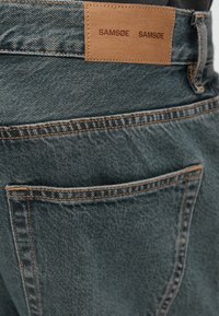 Vaqueros de mezclilla azul oscuro con una parche de cuero marrón que presenta la marca "SAMSØE". Bolsillo trasero visible con costuras en contraste.